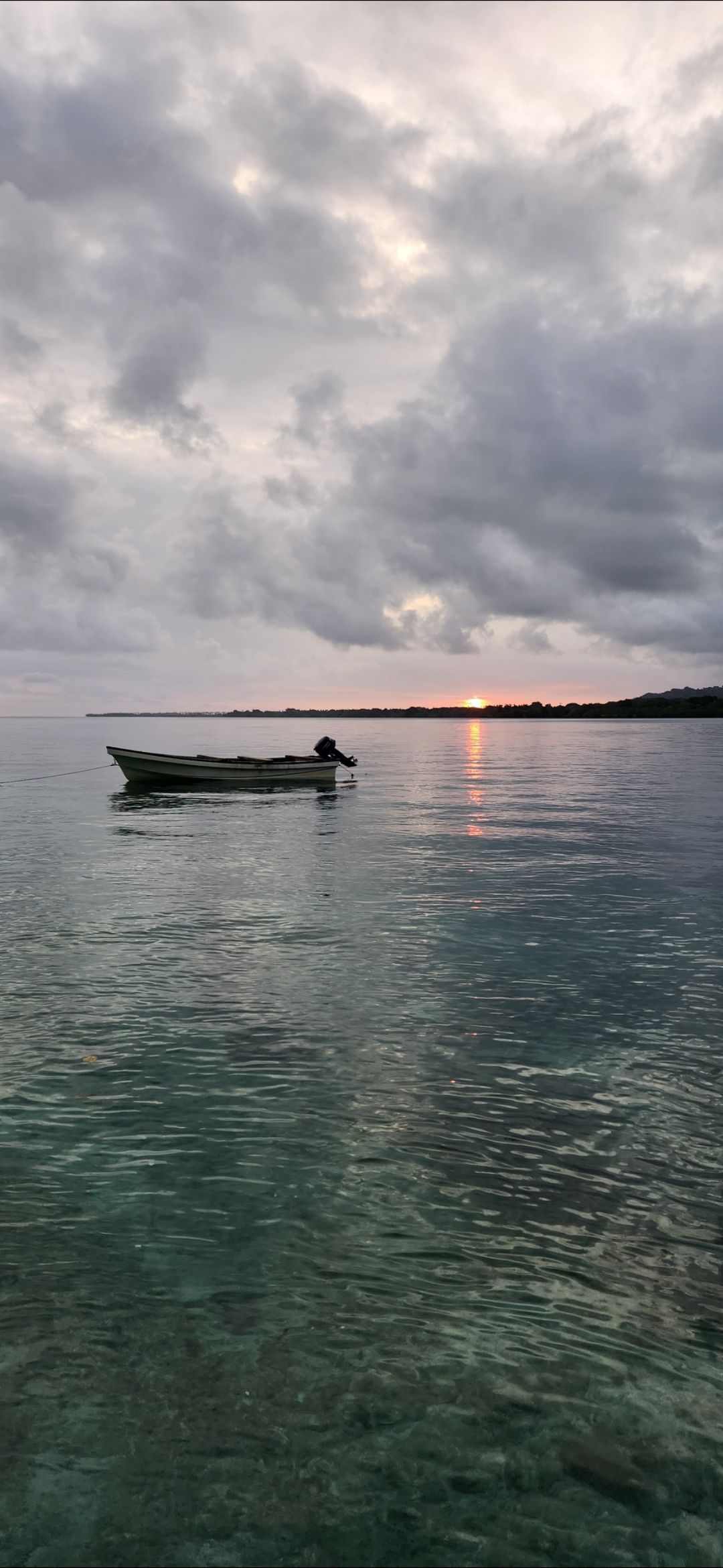 A small boat floats on a calm sea, under a cloudy sky as the sun sets in the distance.