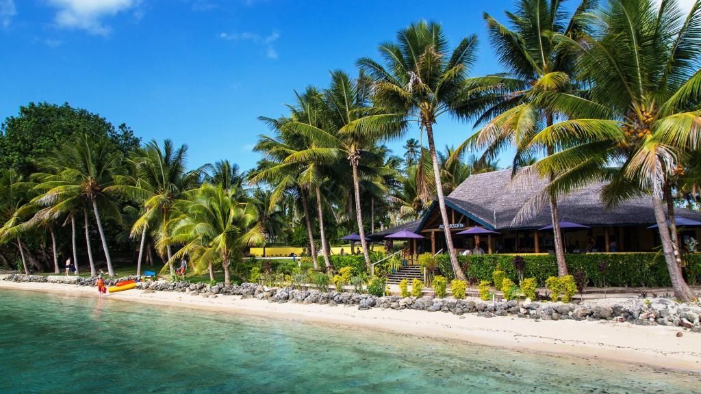 Beachfront resort with palm trees, thatched roof, turquoise water, and clear blue sky.