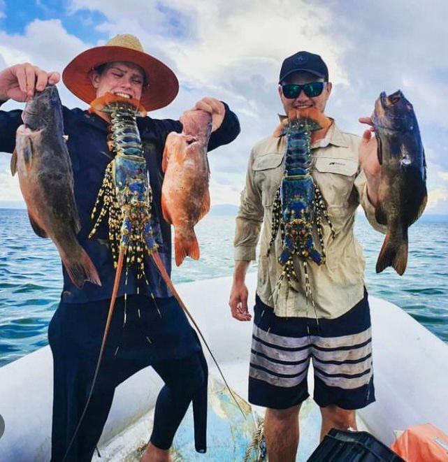 Two men on a boat holding lobsters and fish