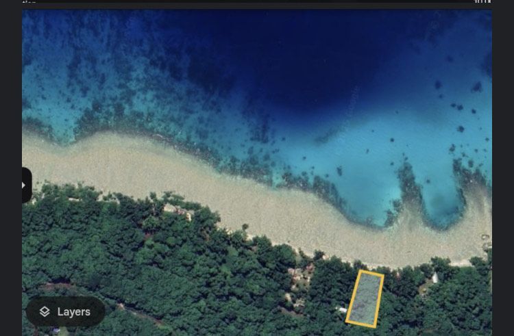 Aerial view of a beach with turquoise water, coral reef, and lush green forest. A rectangular structure is on the land.