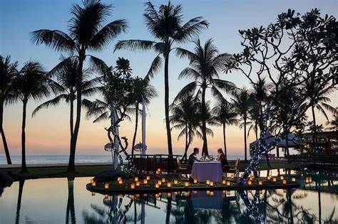 A couple is sitting at a table in front of a swimming pool surrounded by palm trees.