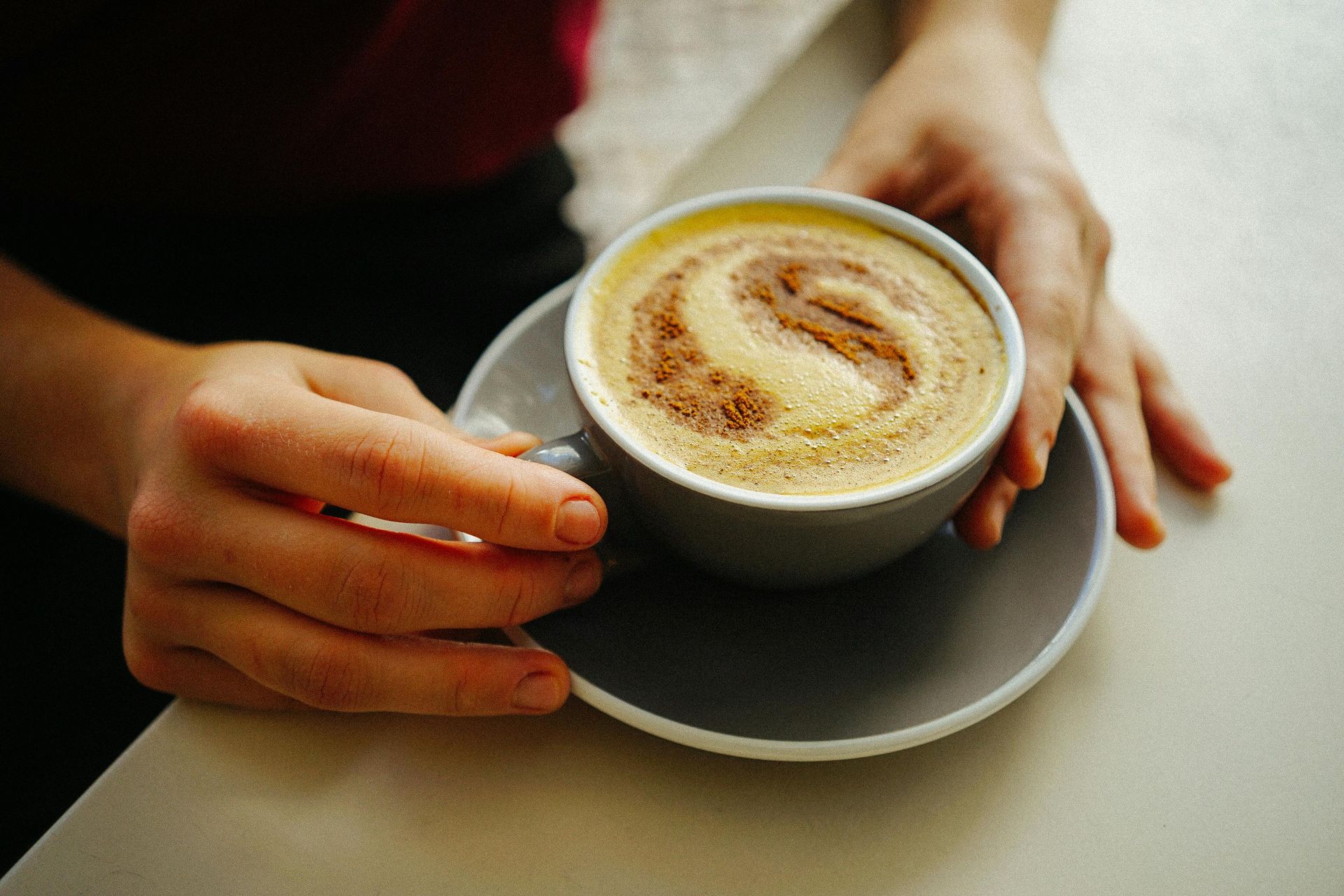 Hands holding a gray mug of cappuccino with a swirl of foam and cinnamon on a matching saucer.