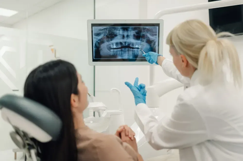 Tandlæge peger på tænder på patientens røntgenbillede vist på en skærm. Patienten sidder i en tandlægestol.