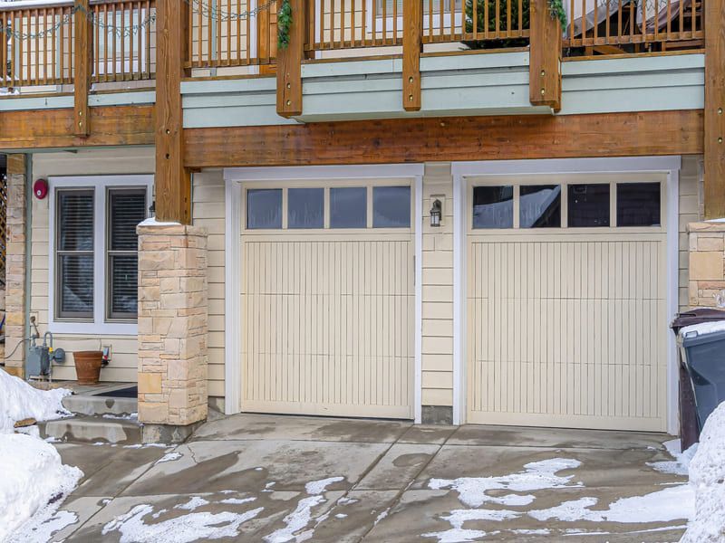Two tan garage doors below a wooden deck; snow on driveway.
