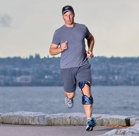 A person in a gray t-shirt and shorts jogs along a waterfront path, wearing a blue knee brace on their left leg.