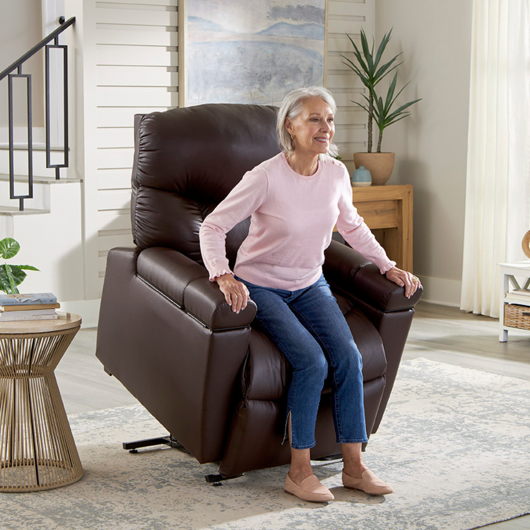 A dark brown leather recliner sits on a patterned area rug in a brightly lit living room near a wooden table and stairs.