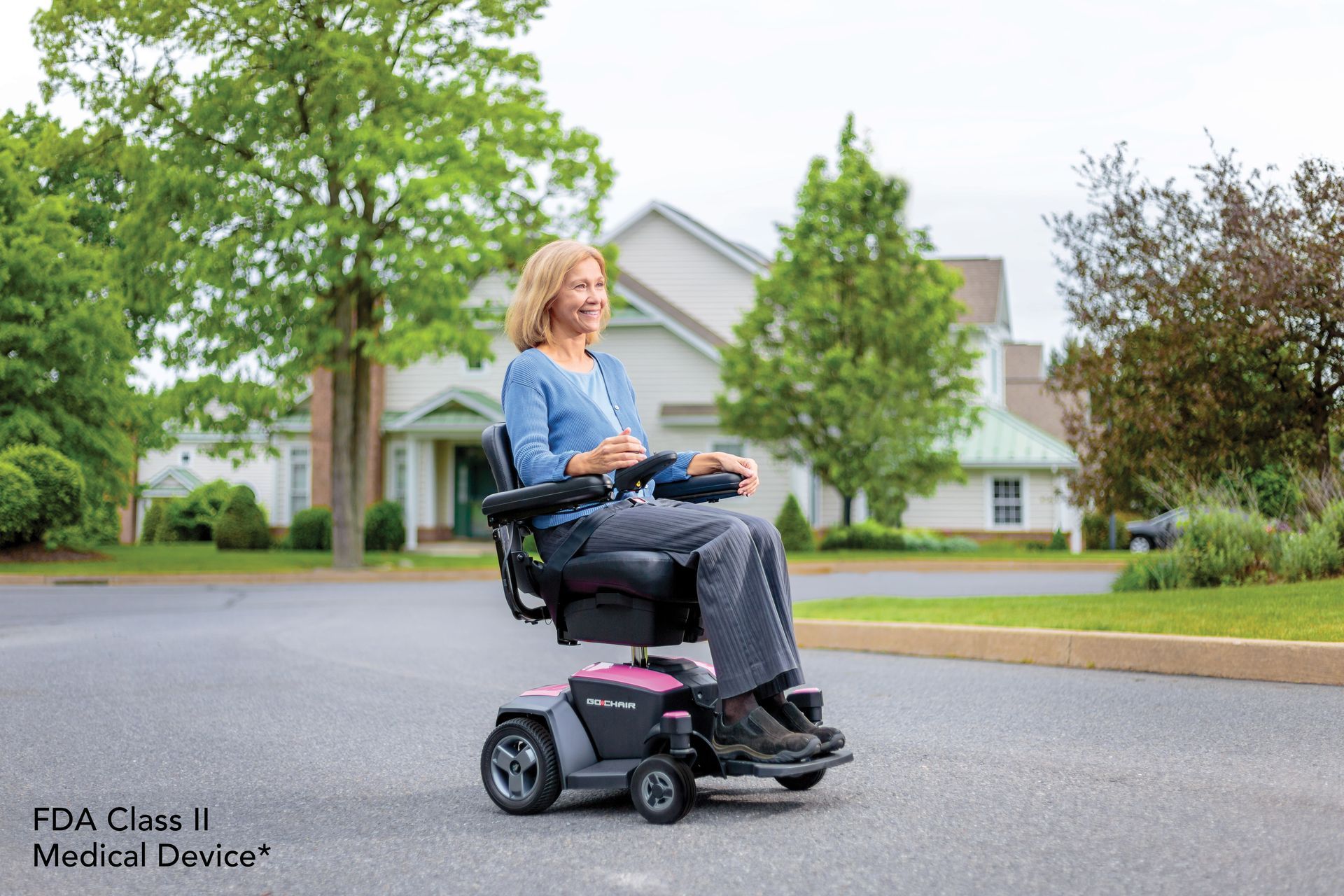 Woman in power wheelchair on a residential street. She is smiling. Houses and trees in background.