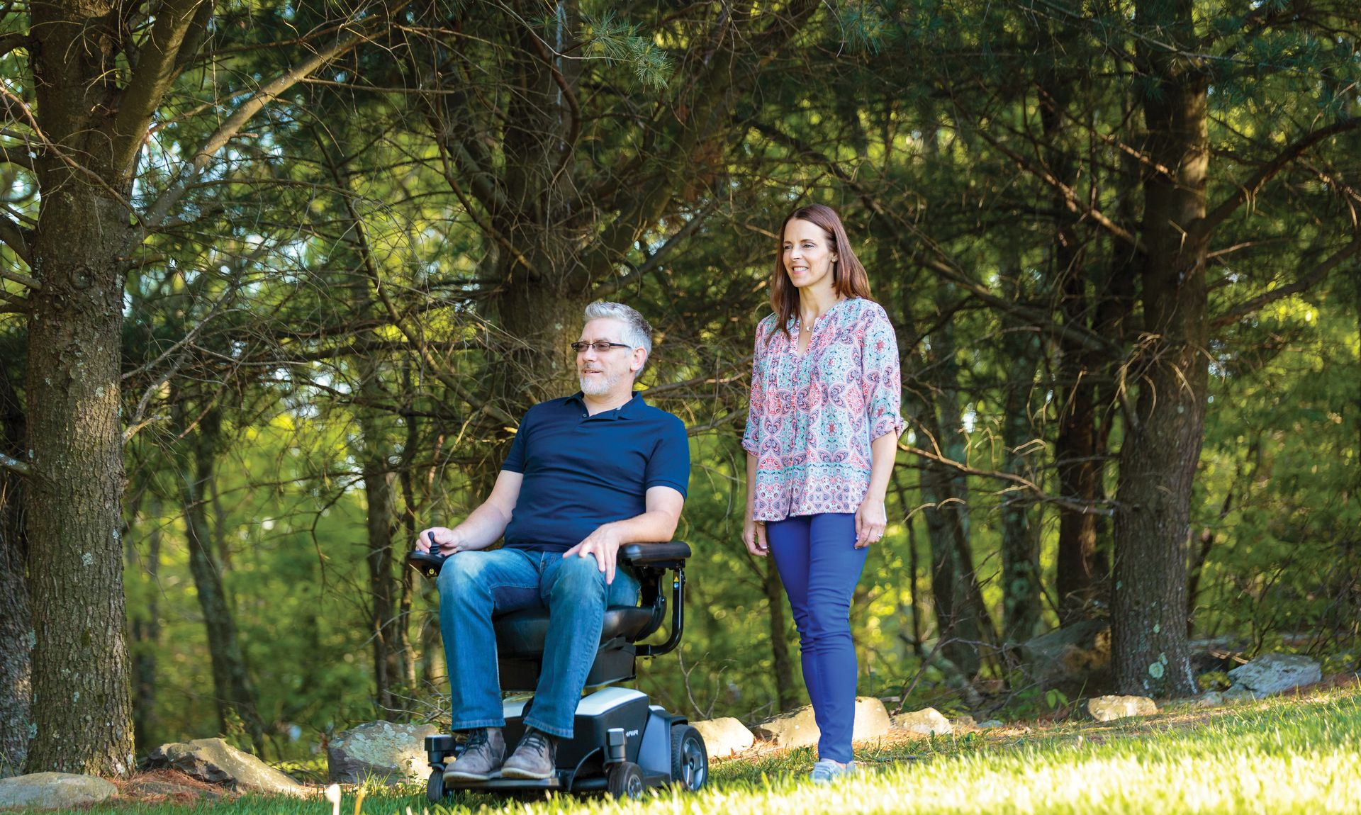 Man in power wheelchair with woman beside him outdoors in grassy area.