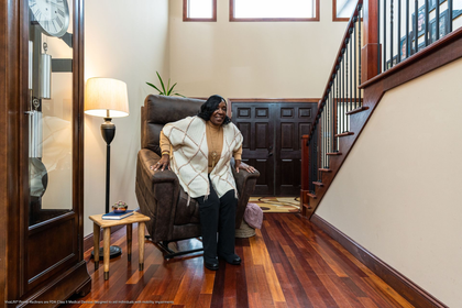 A person sitting on the edge of a recliner in a home entryway with dark wood floors, a stairwell, and a grandfather clock.