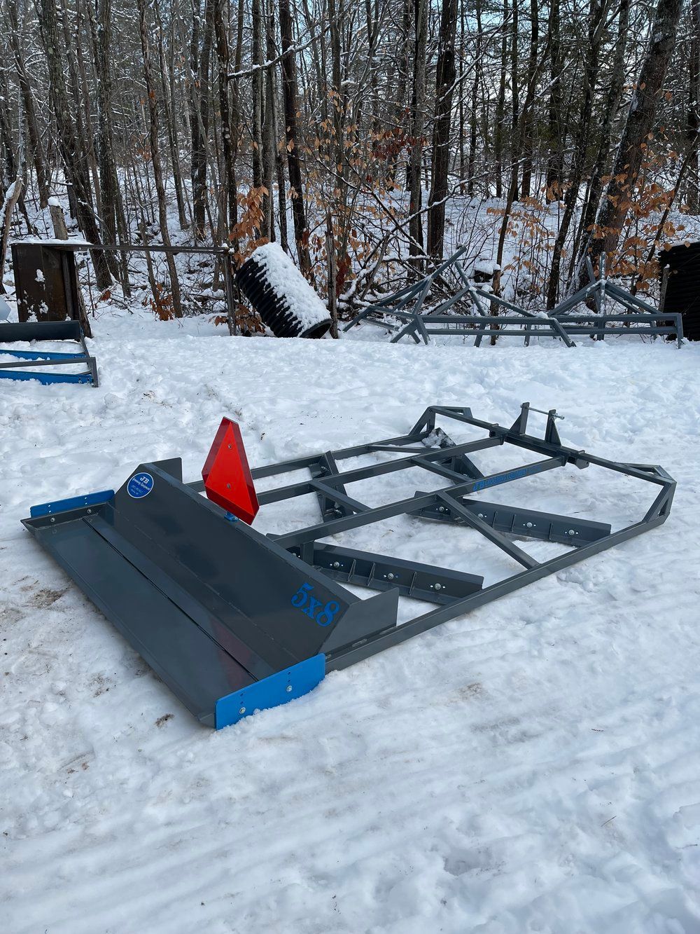 A snow plow is sitting in the snow in front of a forest.