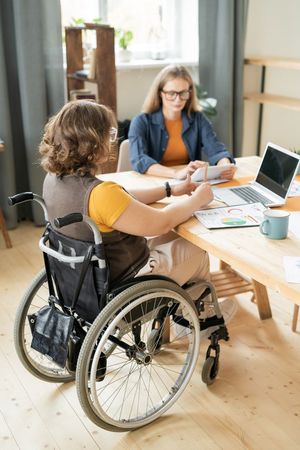Two people collaborate at a table in an office, with one seated in a wheelchair and a laptop open.