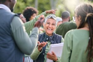 Smiling woman speaking outdoors to a small group, holding papers and gesturing during a discussion