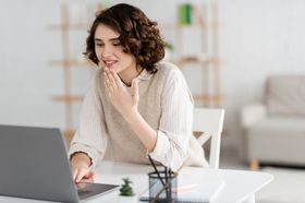 Person smiling at a laptop in a bright office, holding a phone and resting their chin on a hand.