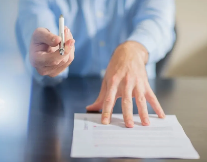 A man is sitting at a table holding a pen and writing on a piece of paper.