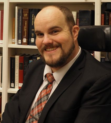 A man in a white shirt and tie is smiling in front of a bookshelf filled with books.