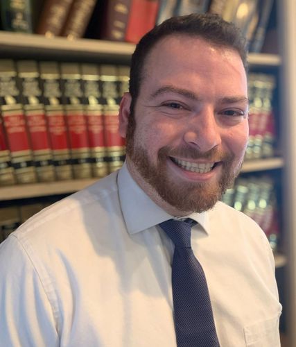 A man in a white shirt and tie is smiling in front of a bookshelf filled with books.