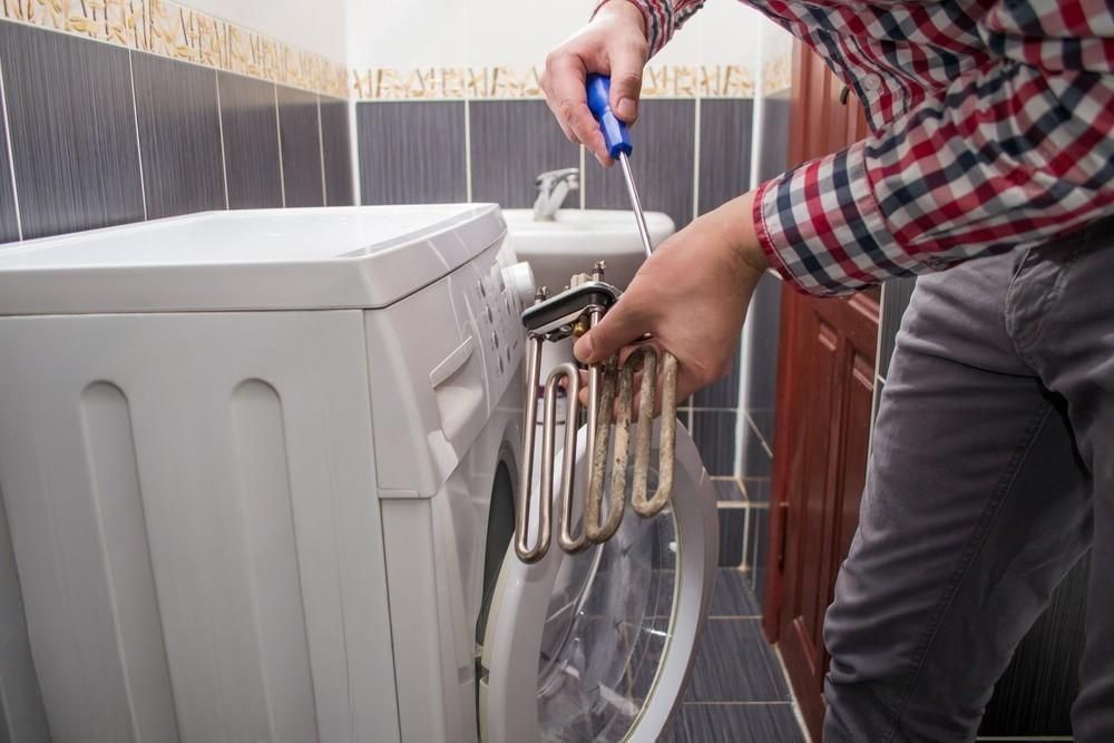 A man is fixing a washing machine with a screwdriver in a bathroom.