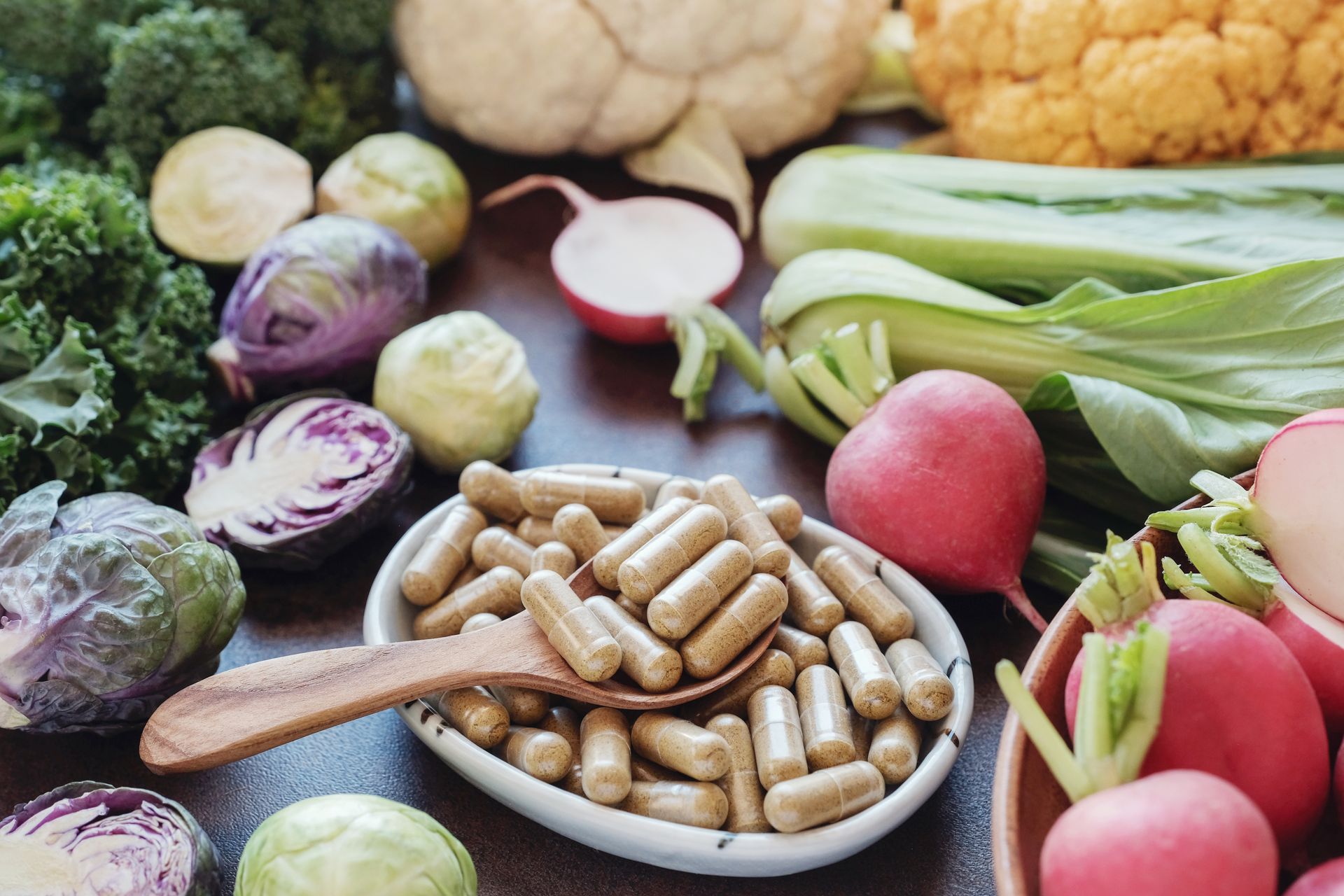 Vegetables (kale, cabbage, radishes) surround a bowl of brown capsules.