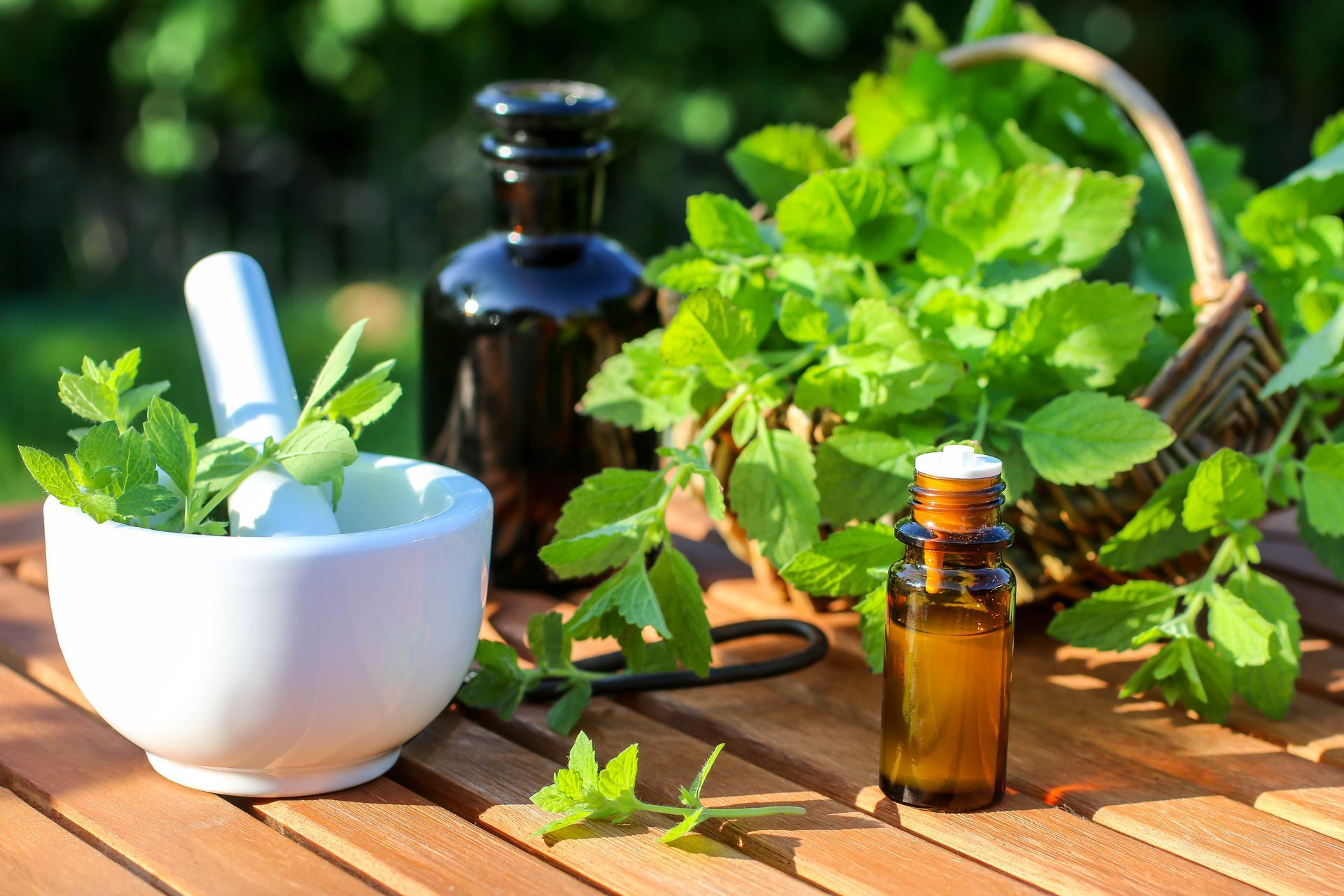 Mortar and pestle with herbs, small bottles of oil, and basket of greenery on a wooden table outdoors.