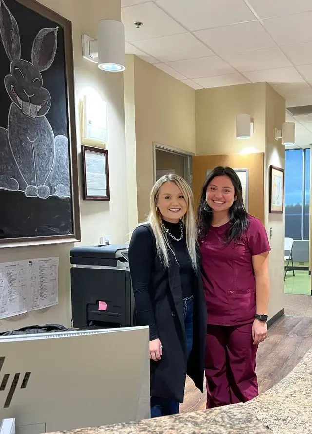 Two people stand near a reception desk in an office. One in a black coat, the other in maroon scrubs, pose for the camera.