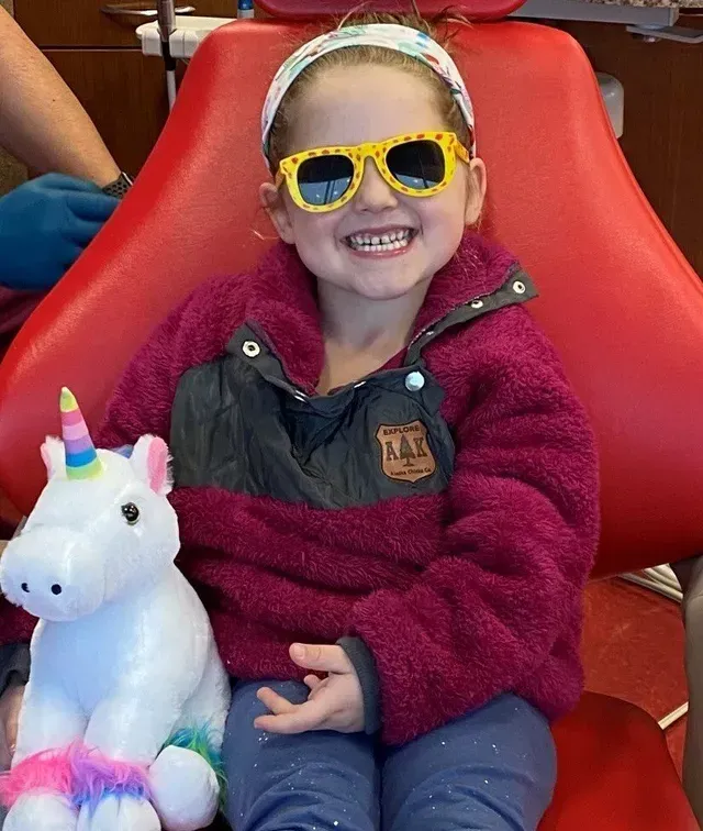 Young girl in sunglasses and a pink fleece smiles with a unicorn toy in a dentist's chair.