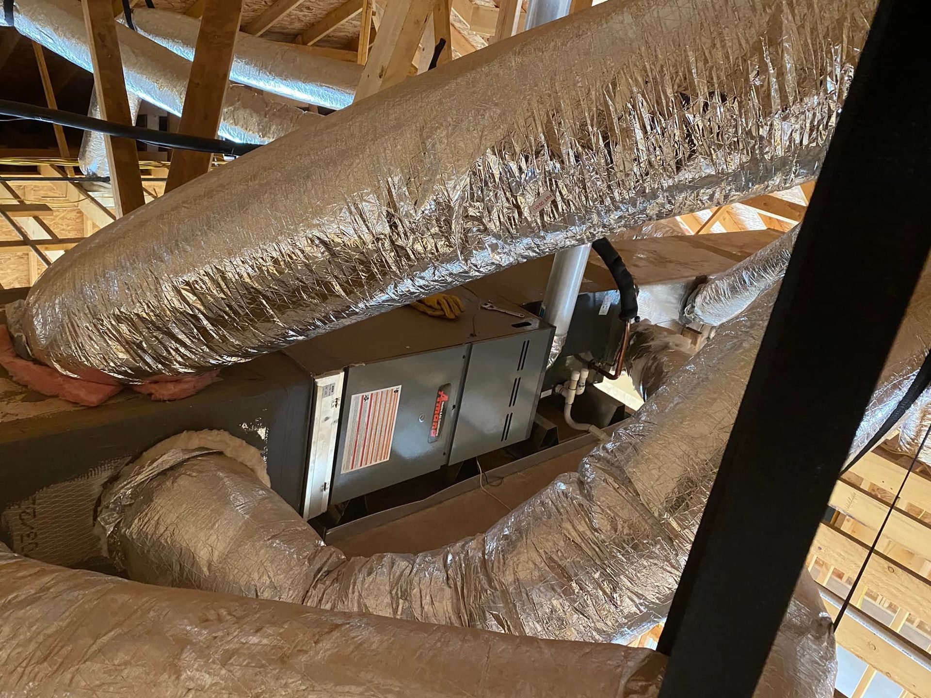 HVAC unit in attic surrounded by insulated ductwork and wooden rafters.