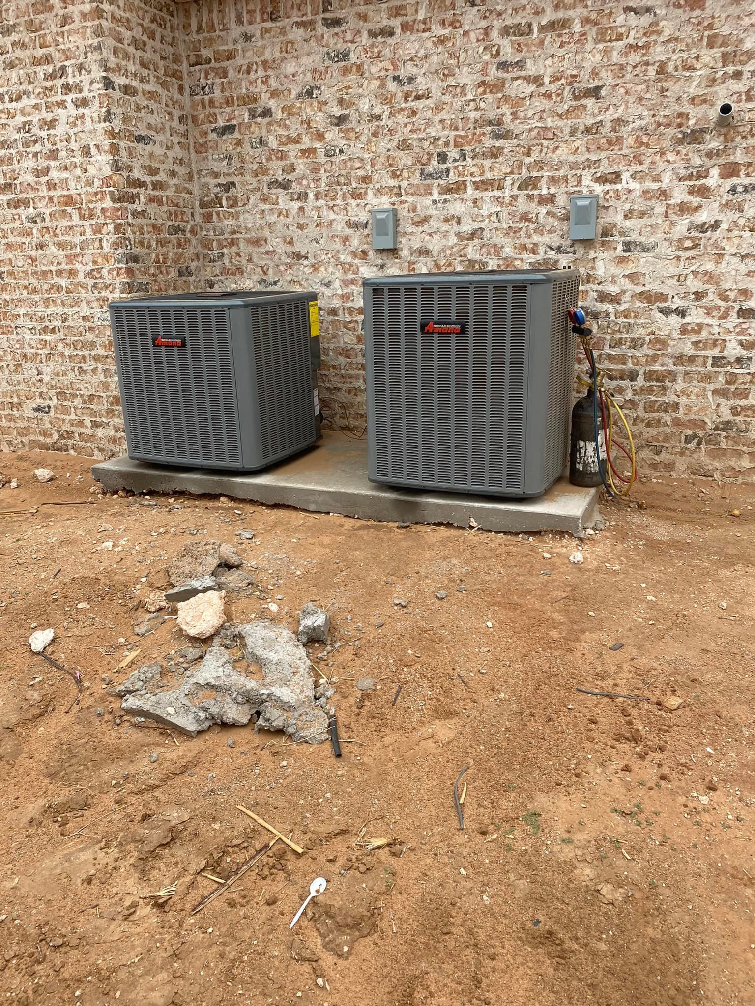 Two gray air conditioning units on a concrete pad, against a brick wall, in a dirt yard.