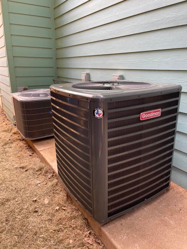 Two Goodman air conditioning units beside a teal siding wall on a concrete pad.