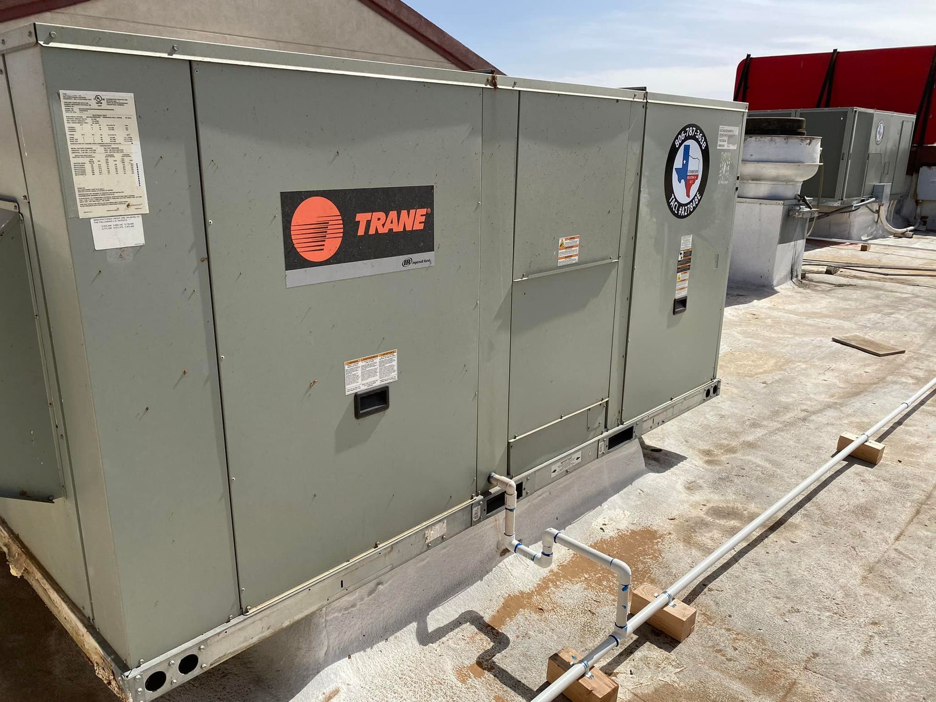 Gray Trane rooftop HVAC unit on a flat, white roof. Piping and other units visible.