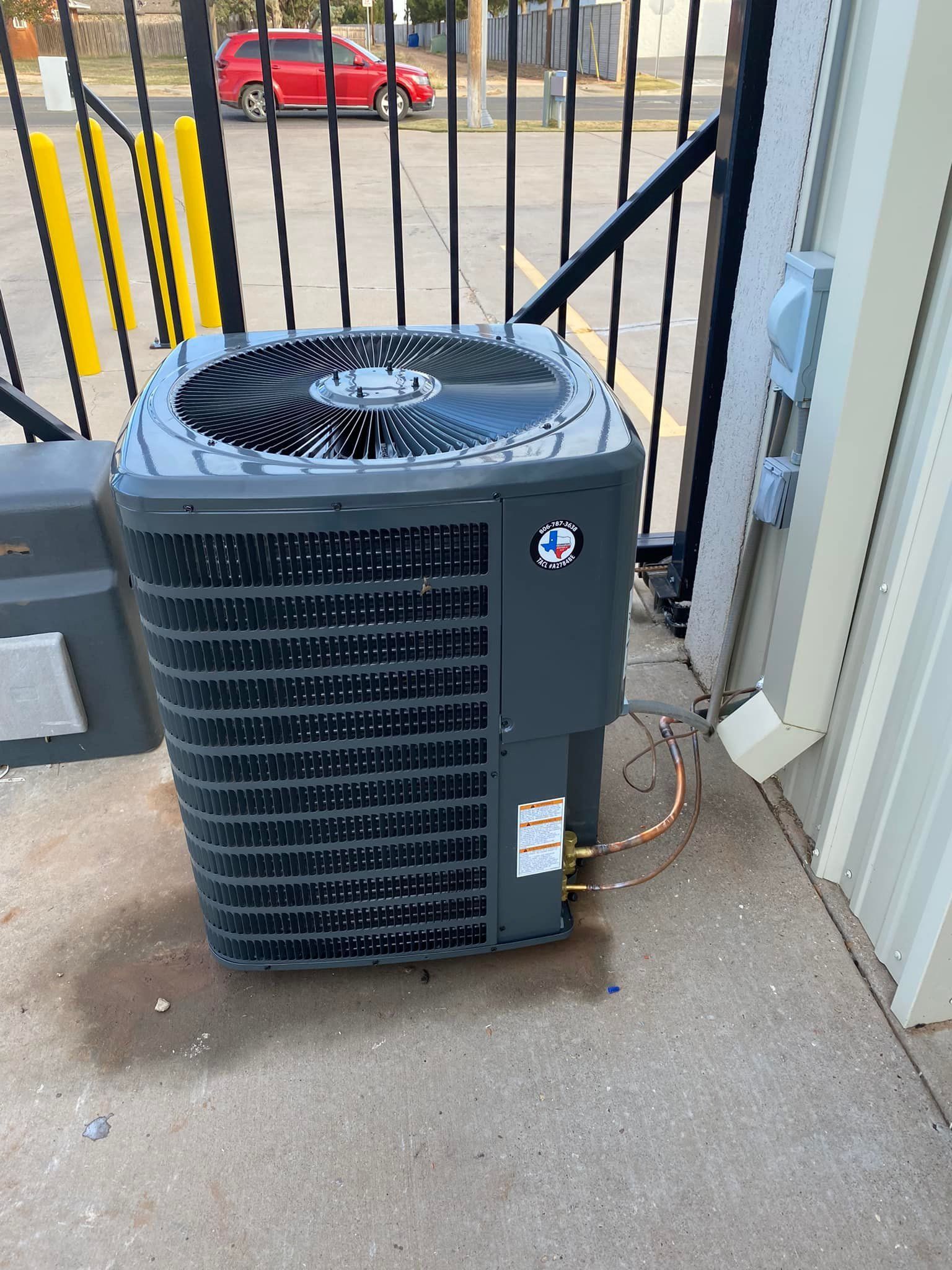 Gray air conditioning unit outside, near a gate and building.