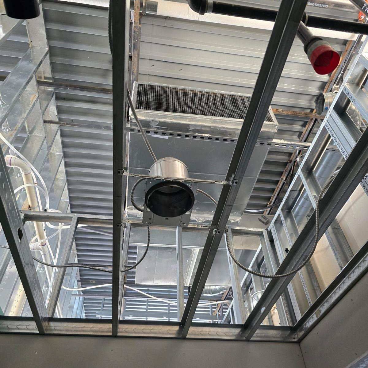 Metal framework of a construction site ceiling. A ventilation fan is visible with exposed wiring and pipes.