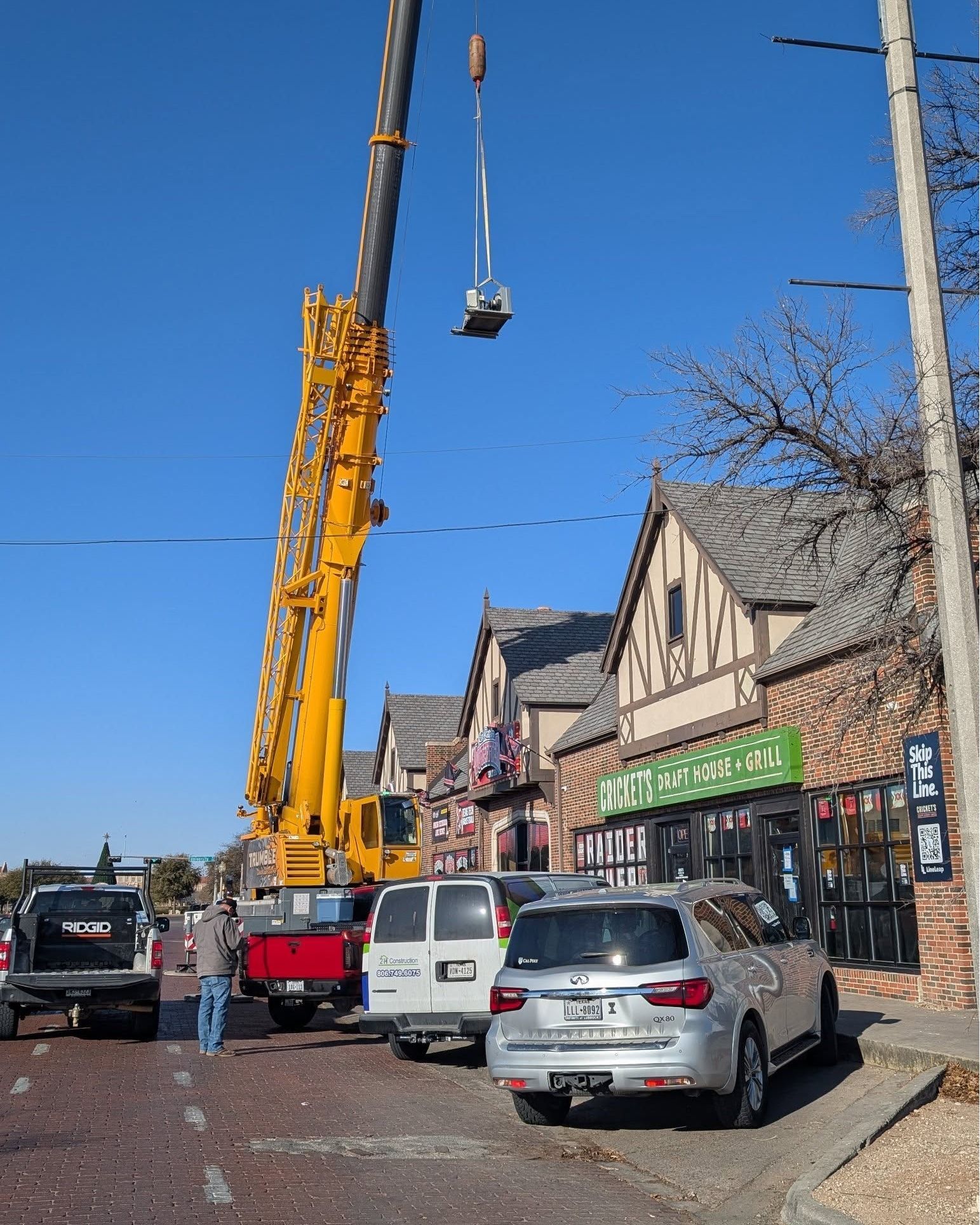A crane lifting a square metal object near brick buildings and parked vehicles on a sunny day.