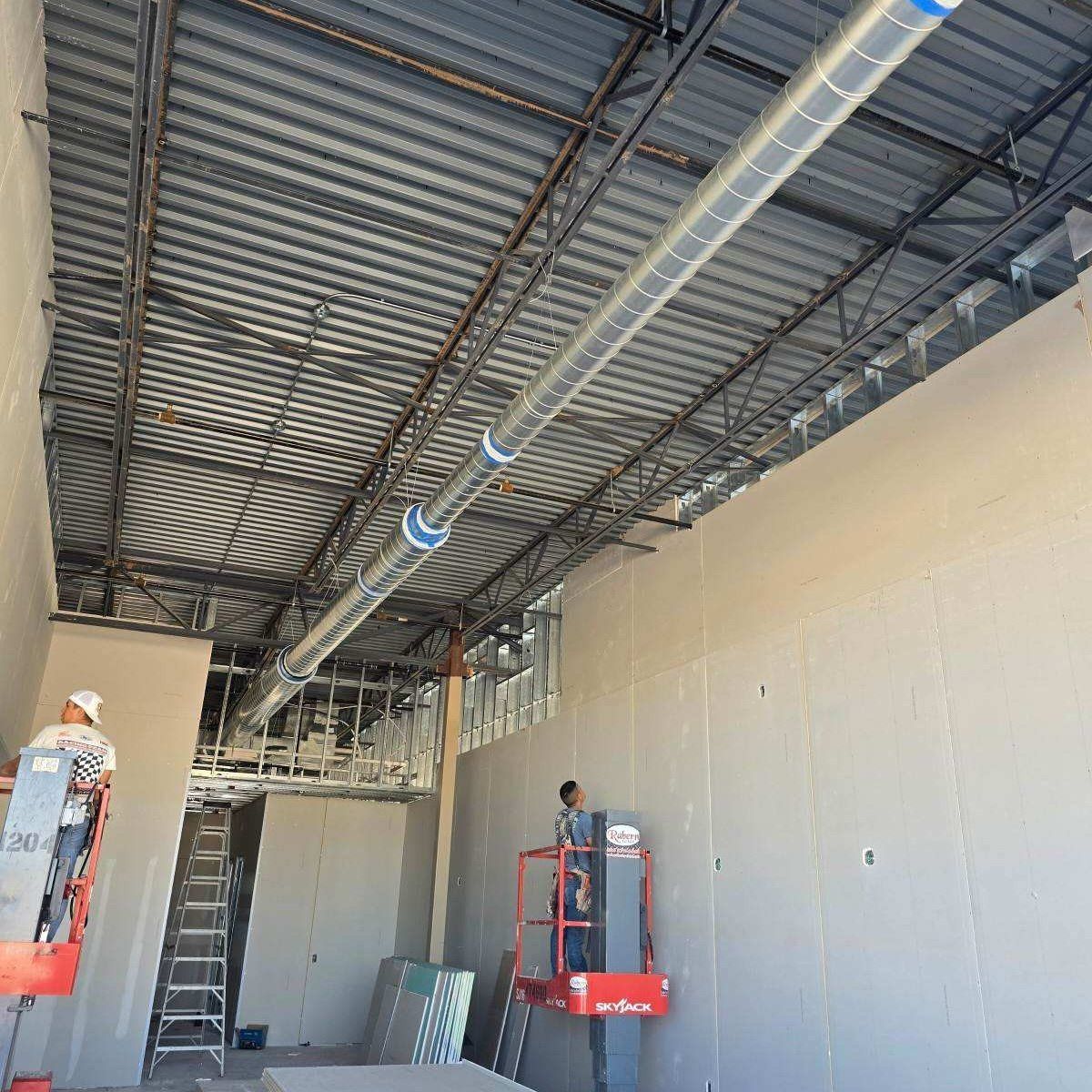 Construction site interior with exposed ceiling beams, HVAC ducting, and workers on lifts.