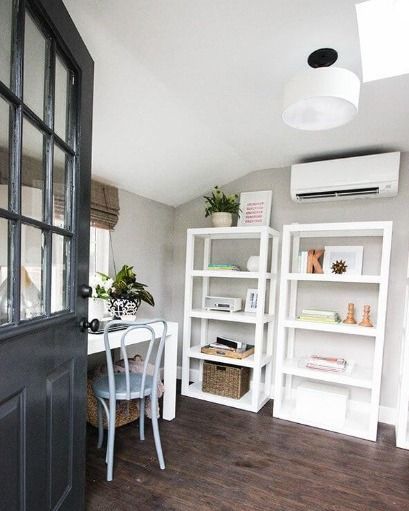 Home office with white shelves, desk, and dark gray door. Wooden floor, neutral walls, and a wall-mounted air conditioner.