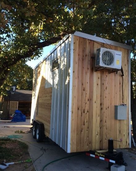Tiny house on wheels with wood siding, metal roof, air conditioner. Parked outdoors.