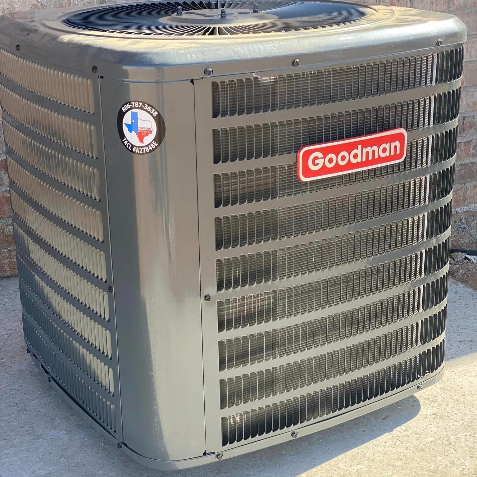 Goodman air conditioner unit outside against a brick wall. Gray metal with a black fan on top.