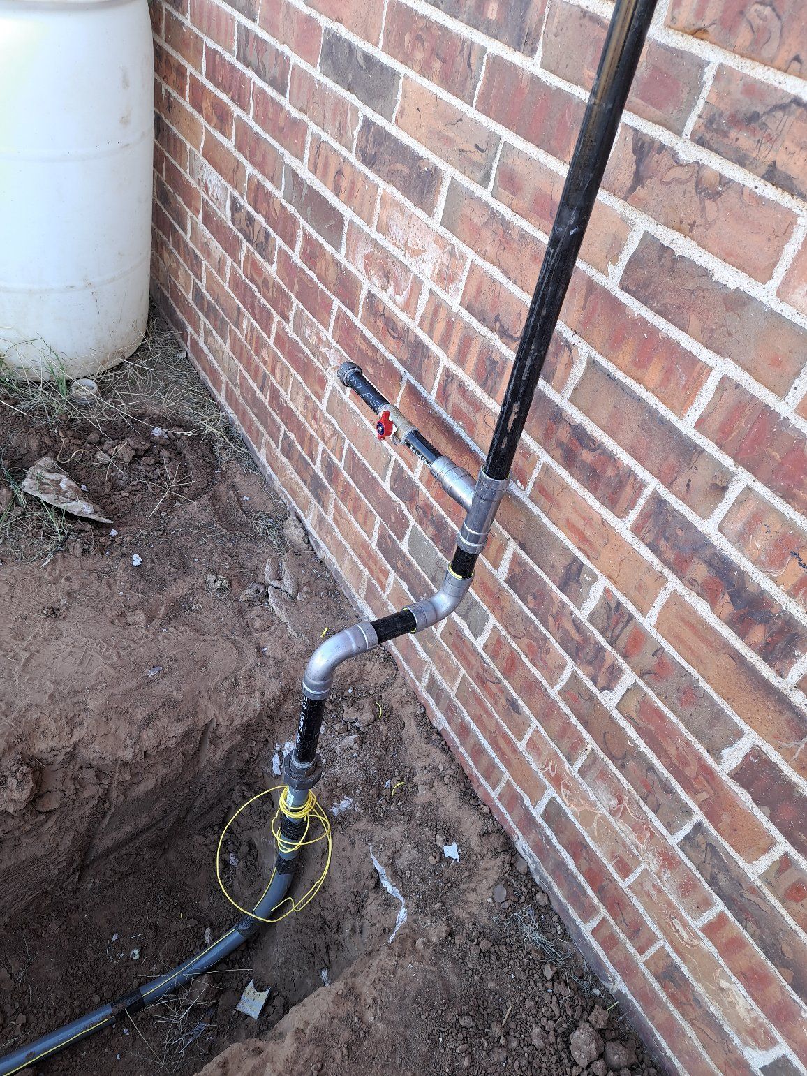 Plumbing with black pipes and a valve against a brick wall. A white barrel is in the background.