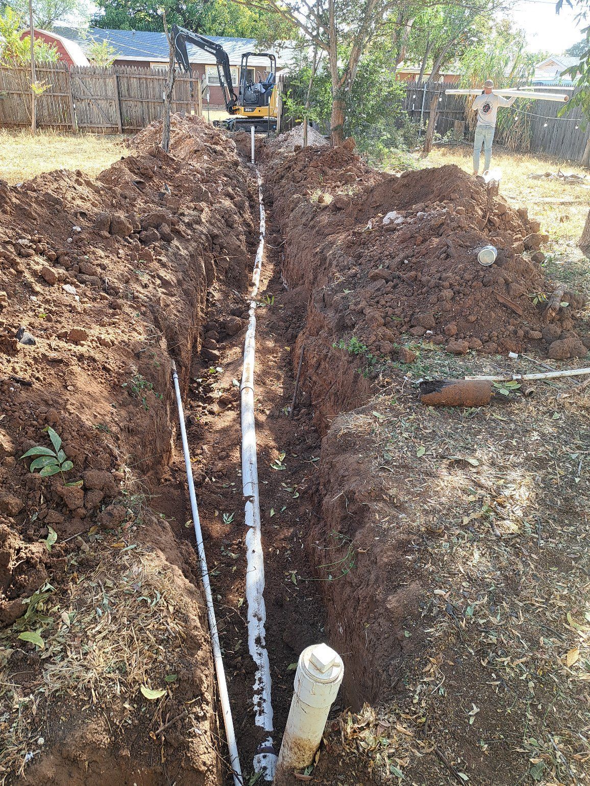 A trench with pipes is being dug in a yard, with an excavator in the background.