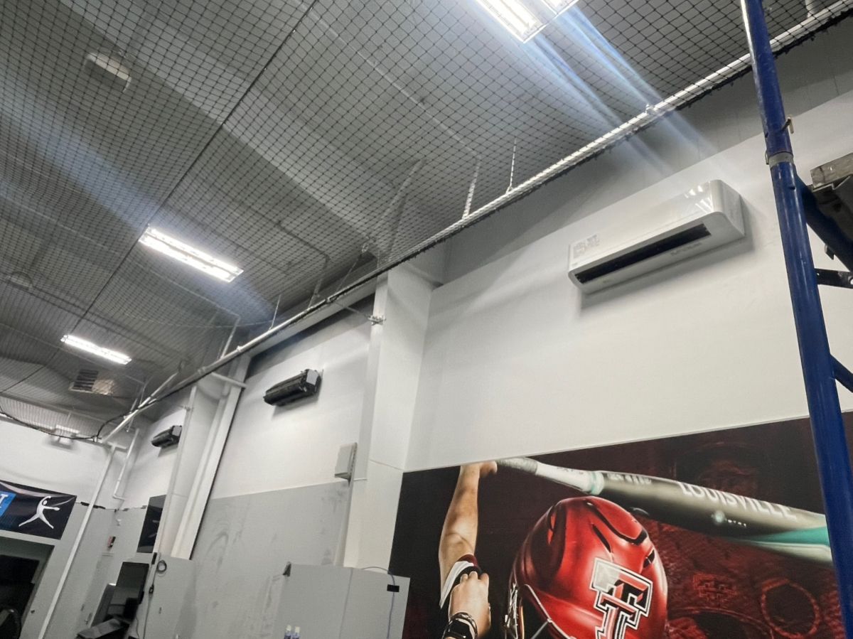 Indoor baseball training facility with protective netting, AC unit, and baseball-themed artwork.