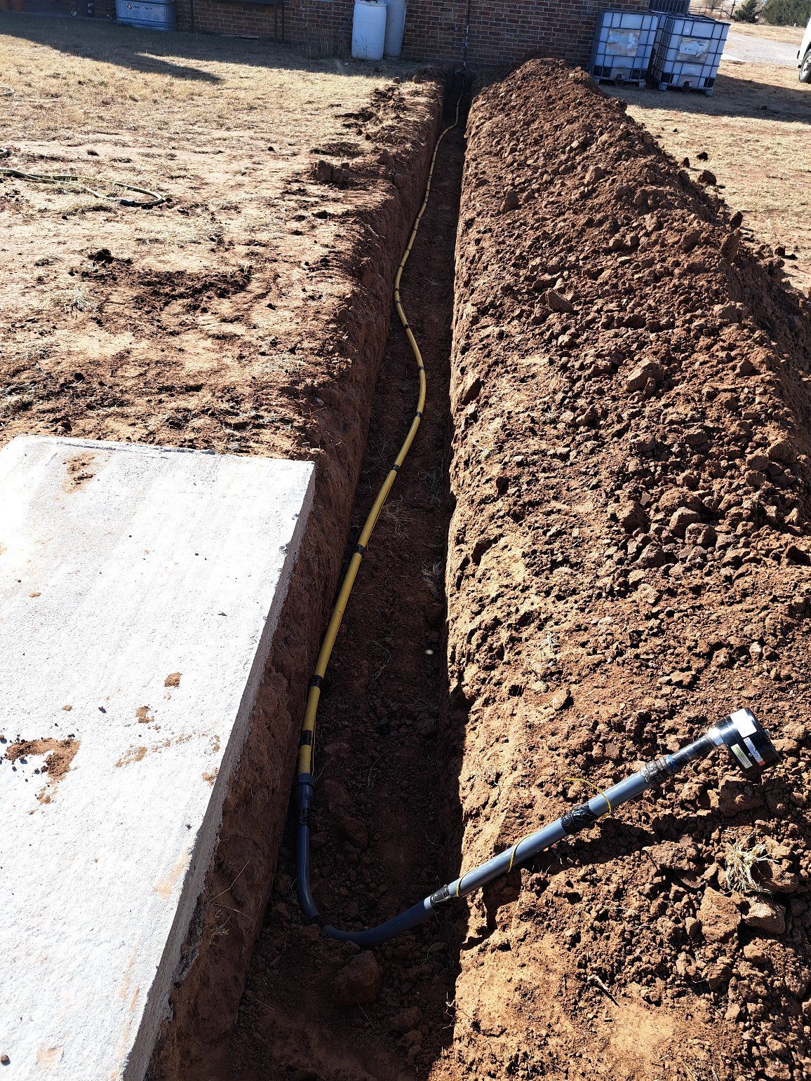 A trench dug in dirt, containing yellow conduit and a black pipe, possibly for utilities.
