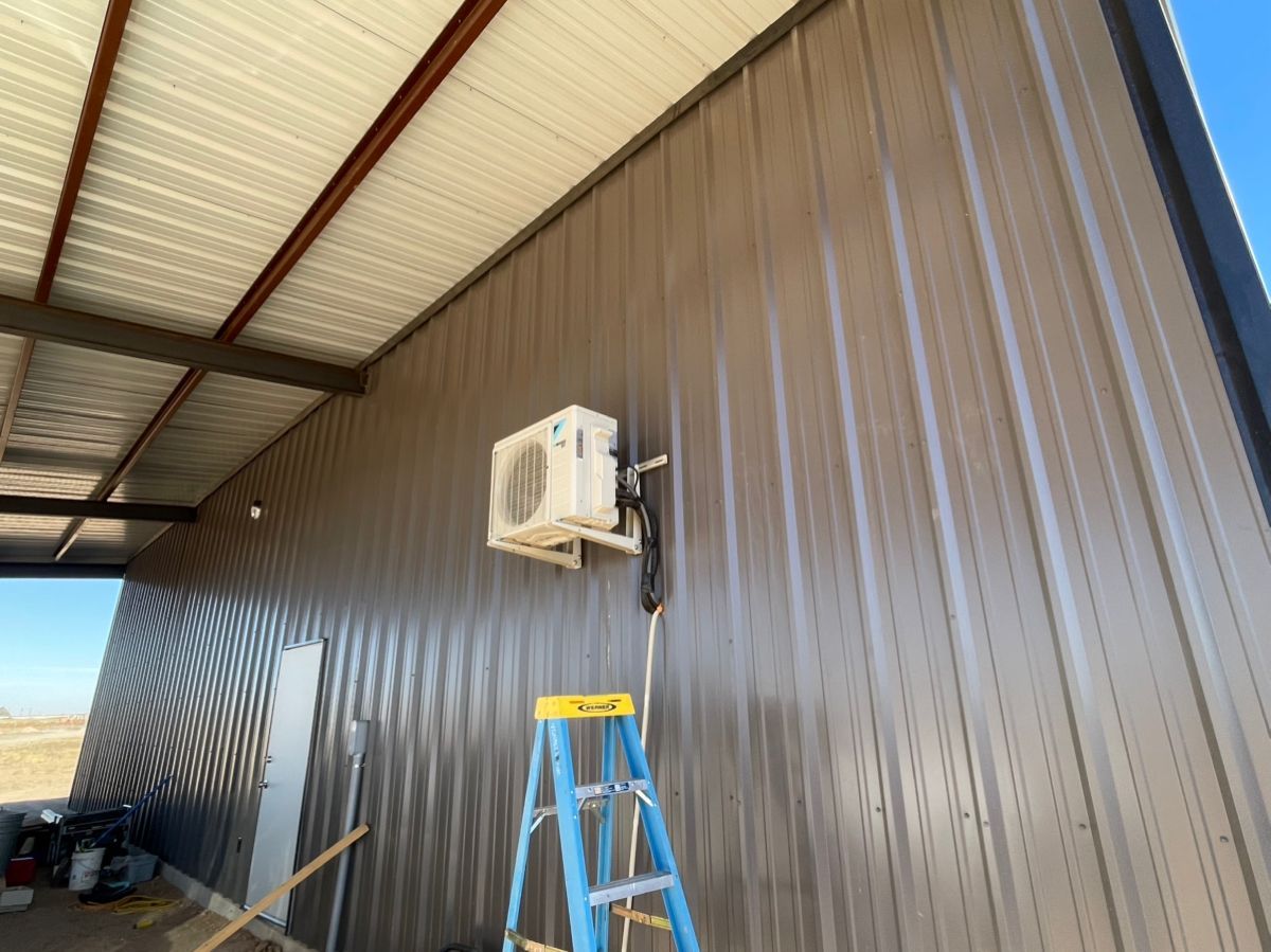 Air conditioner unit mounted on a metal-sided building wall, with a ladder and awning visible.