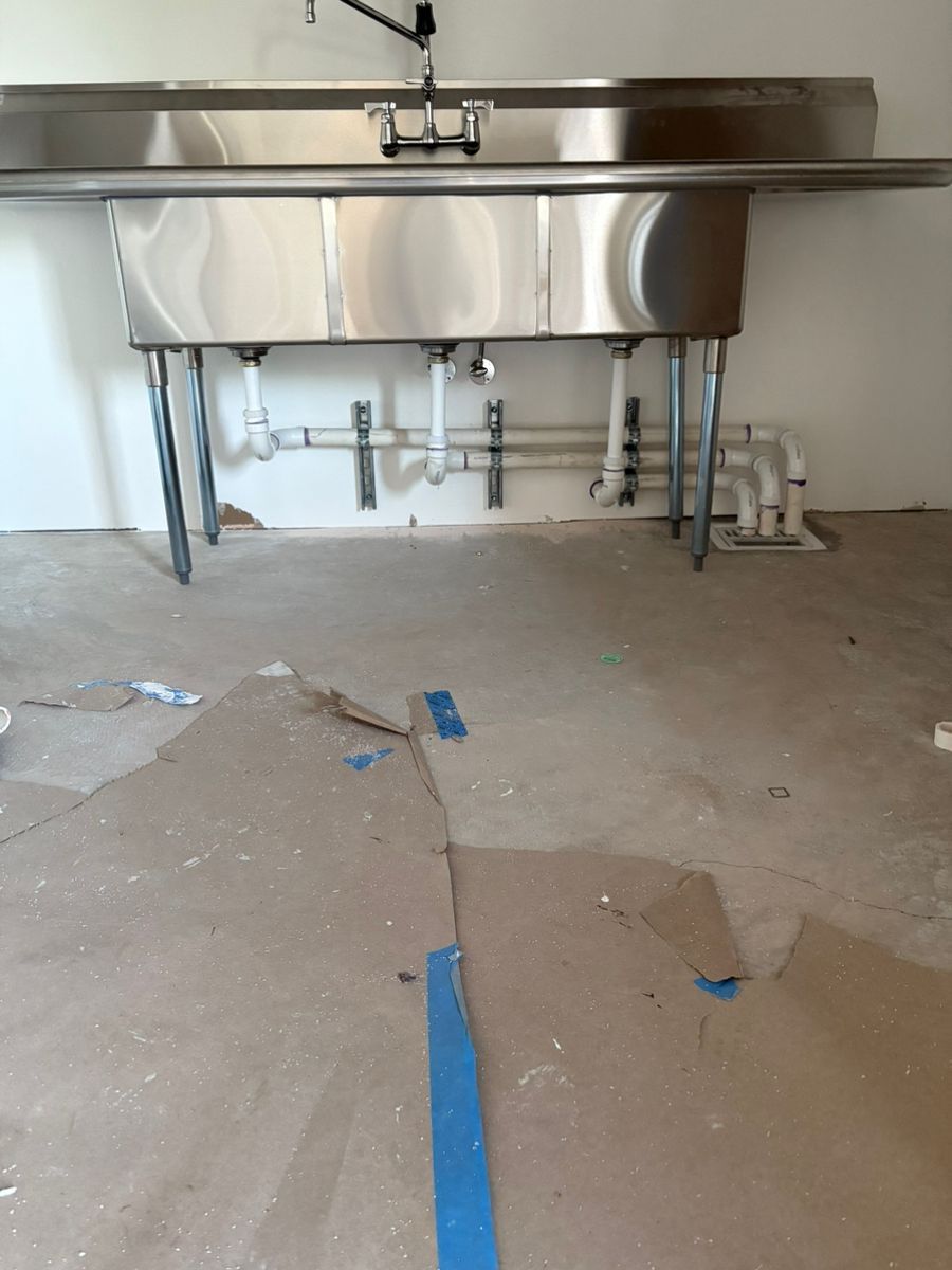 Stainless steel triple sink in a room with exposed pipes and cardboard on the floor.