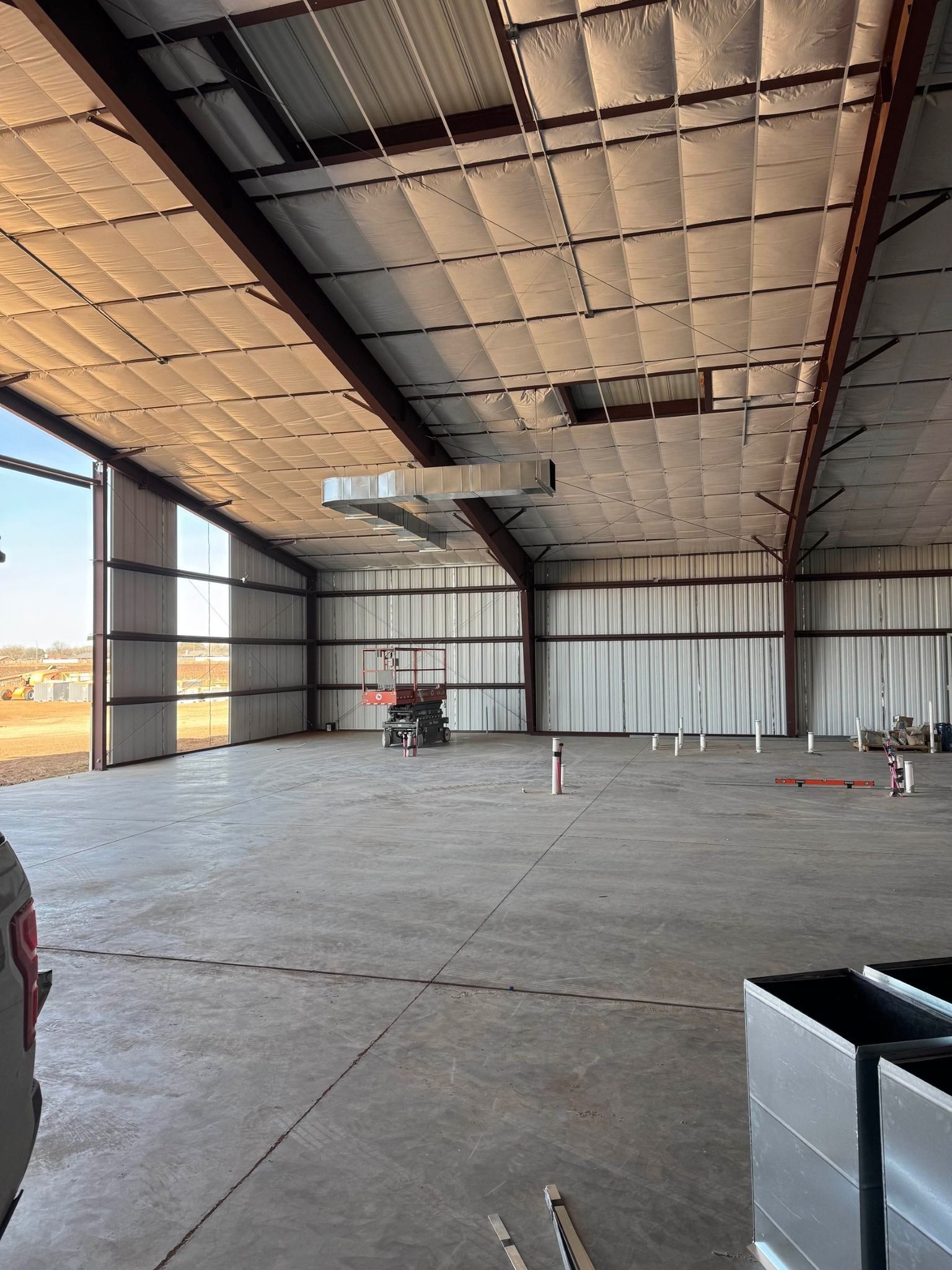 Interior of a large metal warehouse with open door, showing concrete floor, insulation, and machinery.