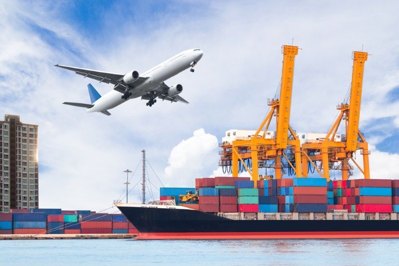 A plane is flying over a cargo ship in the water.