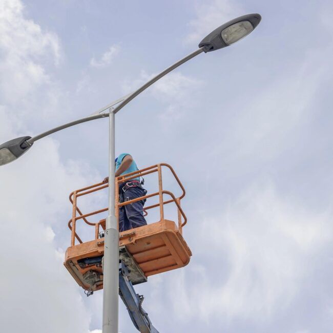 Person in an orange lift platform repairs a street light under a cloudy sky.