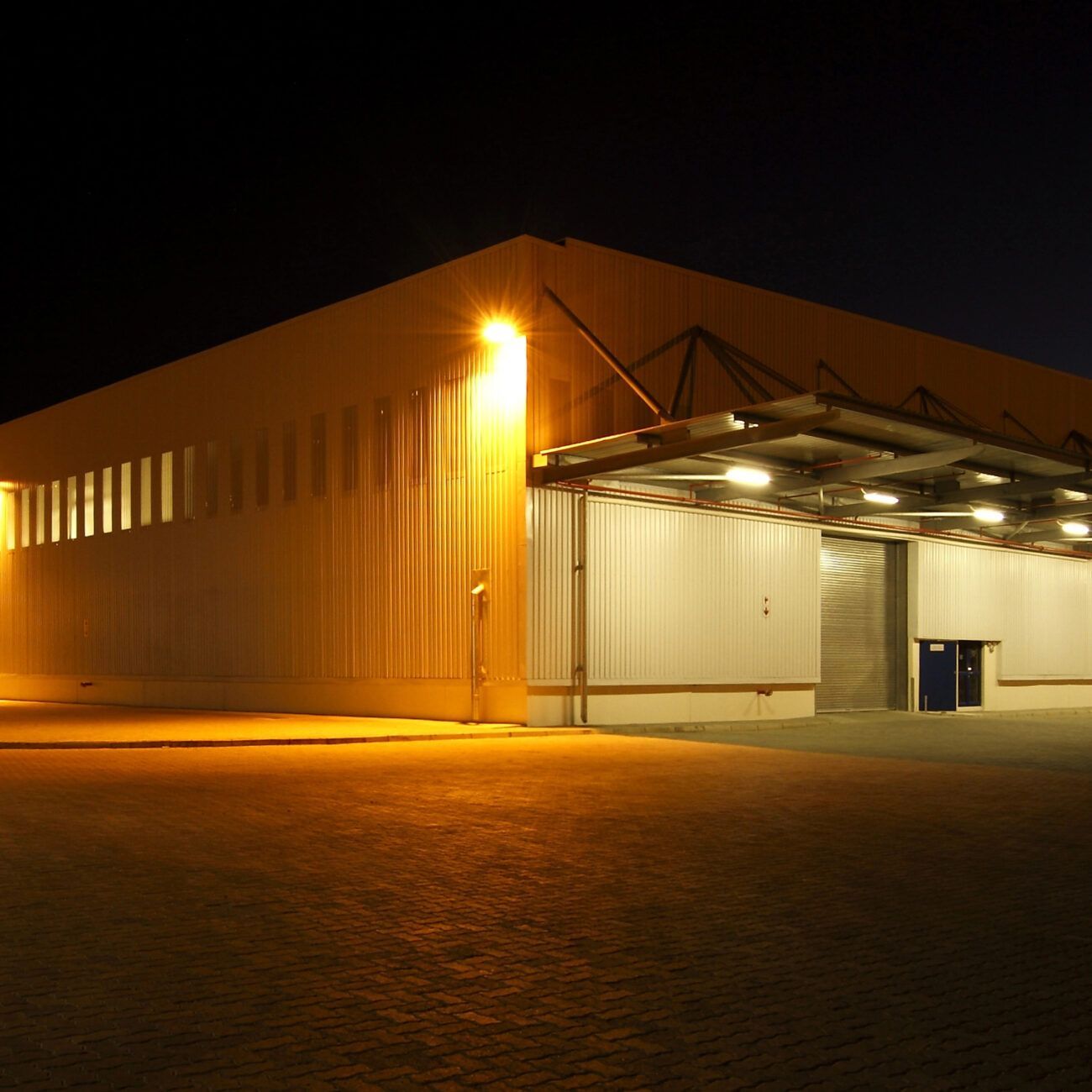 Warehouse exterior at night, lit by bright orange lights. Corrugated metal siding and loading bays are visible.