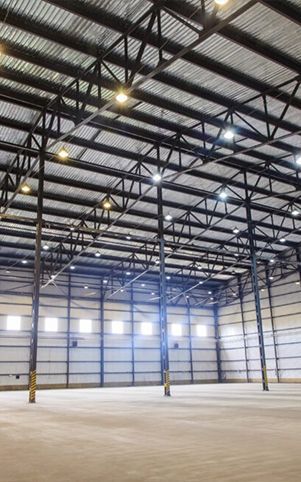 Interior of a large, empty warehouse with a metal beam ceiling and concrete floor, lit by overhead lights.