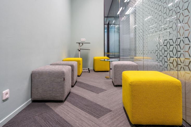 Small modern office space with yellow and gray cube seats, gray patterned carpet, glass wall.