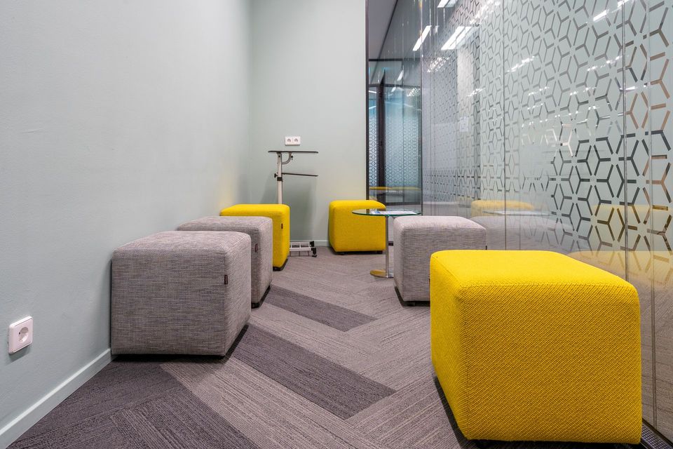 Small modern office space with yellow and gray cube seats, gray patterned carpet, glass wall.
