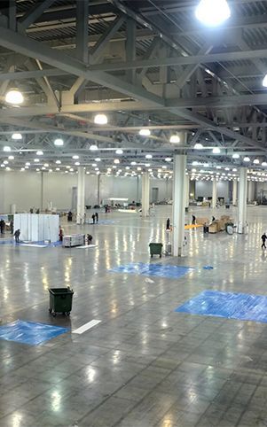 Large, empty convention hall with high ceiling and bright lights. Several workers are present, along with trash cans and blue floor markings.